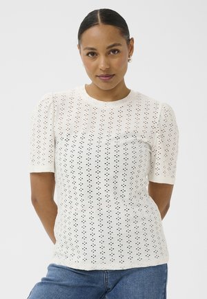 Femme aux cheveux foncés coiffés en chignon bas, portant des boucles d'oreilles créoles et un haut blanc à manches courtes en broderie anglaise avec un col rond, associé à un jean bleu.