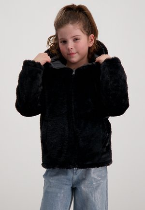 Zwarte nepbont jas met ritssluiting aan de voorkant, grote capuchon en zijzakken. Zachte textuur en losse pasvorm. Gepaard met lichtblauwe spijkerbroeken.