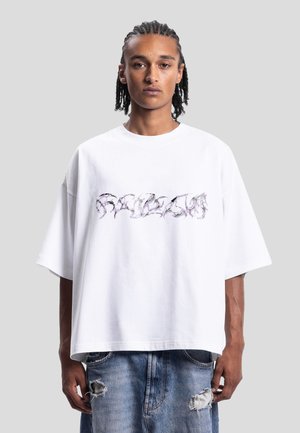 Ung mann med flettet hår iført en oversized hvit T-skjorte med abstrakt svart og lilla trykk og slitte blå jeans, stående rett opp.