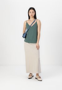 Green scalloped neckline top, beige ribbed maxi skirt, and beige slip-on shoes with black soles; model carrying small blue crossbody bag.