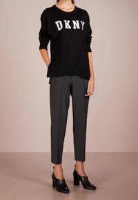 Žena v černém svetru DKNY, šedých zužujících se kalhotách a černých botách na podpatku stojí před jednobarevným pozadím.