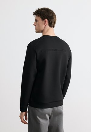 Young man facing left wearing a black long-sleeve sweatshirt with subtle embossed text on the upper back and gray pants.