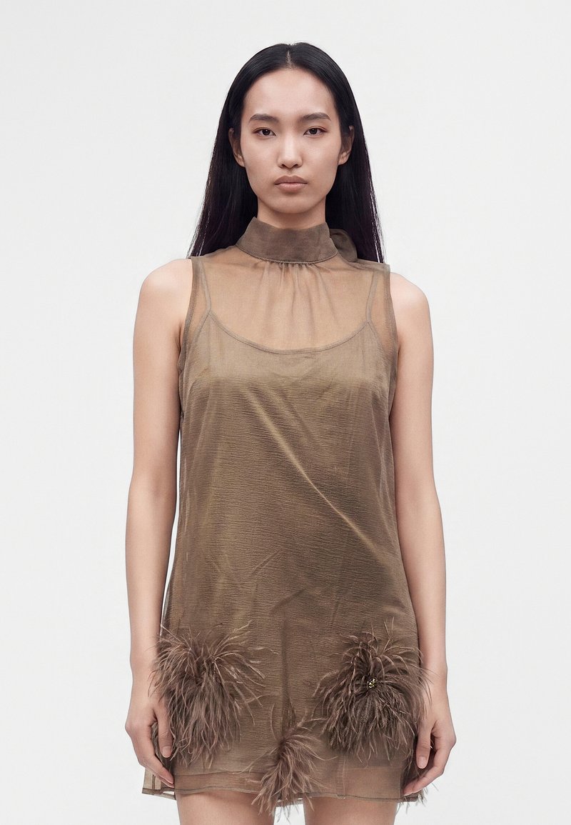 Jeune femme aux longs cheveux foncés vêtue d'une robe marron transparente sans manches avec des détails à plumes près de l'ourlet, debout devant un fond uni.