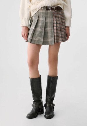 Femme portant une mini-jupe plissée avec des motifs à carreaux mélangés, des bottes noires montantes et un pull crème avec des détails effilochés.