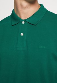 Polo en coton vert avec un col classique, deux boutons nacrés et un logo "Esprit" brodé sur la poitrine. Tissu à texture lisse.