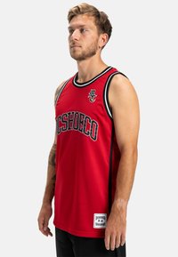 Červená basketbalová dres z oka tkaniny s čiernymi detailmi a výrazným grafickým textom. Bočné panely pridávajú štruktúru. Obsahuje značkovú etiketu.