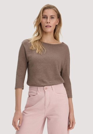 Vrouw met lang blond haar draagt een bruin topje met driekwart mouwen en lichtroze broek met hoge taille tegen een effen achtergrond.