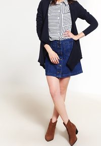Gonna di jeans con bottoni frontali, colore blu scuro, abbinata a una blusa a righe bianche e blu navy e a un cardigan nero. Stivali alla caviglia marroni completano il look.