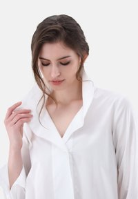 Camicia bianca con un design moderno, caratterizzata da un colletto alto, maniche lunghe e un tessuto liscio e strutturato. Lucentezza leggera e dettagli minimi.