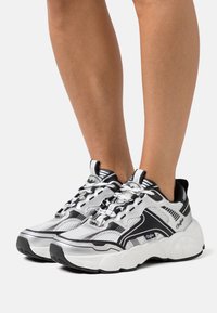 Schwarz-weiße Sportschuhe aus Netz- und Gummi-Materialien, mit einer klobigen Sohle, auffälligen Schnürsenkeln und diagonalen Streifen als Detail.