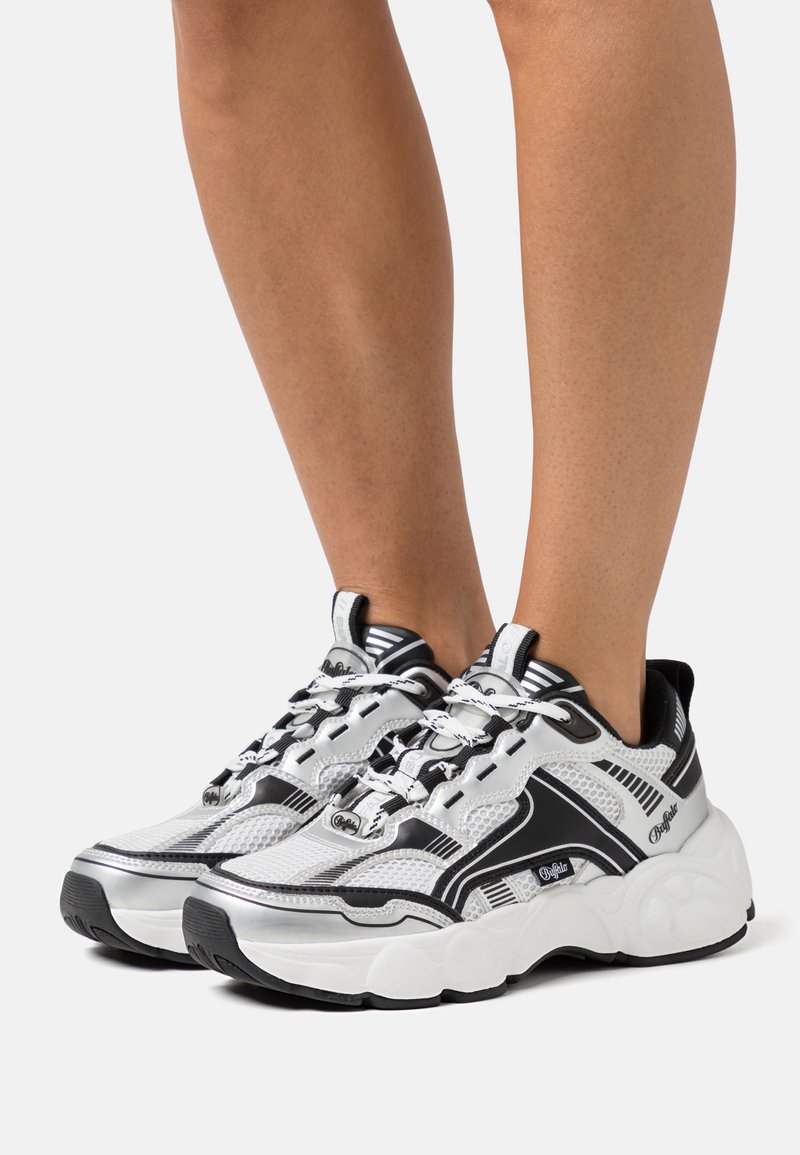 Schwarz-weiße Sportschuhe aus Netz- und Gummi-Materialien, mit einer klobigen Sohle, auffälligen Schnürsenkeln und diagonalen Streifen als Detail.