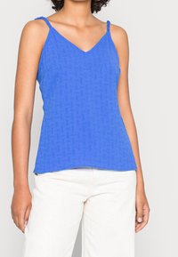 Modrá bezrukávová blůza s výstřihem do V, texturovanou tkaninou a zkroucenými ramínky. Design má volný střih a jemný vzor. Spárováno se světlými kalhotami.