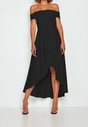 Vrouw draagt een zwarte off-shoulder jurk met een high-low zoom en doorzichtige bandjes op hoge hakken, staand tegen een effen lichte achtergrond.