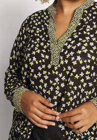 Blouse noire à motif floral blanc et jaune, avec un col en V fendu et des poignets à motifs contrastants. Tissu léger et coupe décontractée.