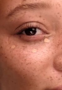 Primo piano di un viso con pelle marrone chiaro. Due piccole macchie beige chiare sono visibili sulla guancia, probabilmente trucco o prodotto per la cura della pelle. Presenti lentiggini.