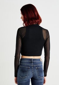 Juodas tinklinis ilgos rankovės crop top'as su aukštu kaklu, derinamas su prigludusiomis tamsiai mėlynomis džinsomis, turinčiomis subtilų galinį kišenės dizainą.