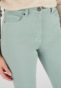 Vaqueros de tiro alto verde claro con una textura suave, que presentan bolsillos frontales y un cierre de botón metálico. Diseño simple y moderno.