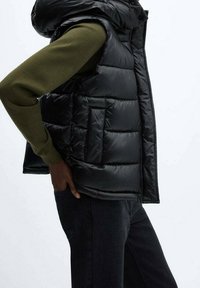 Czarna pikowana kamizelka z kapturem, z przeszyciami, bocznymi kieszeniami i błyszczącym wykończeniem, noszona na zielonej koszuli z długim rękawem.