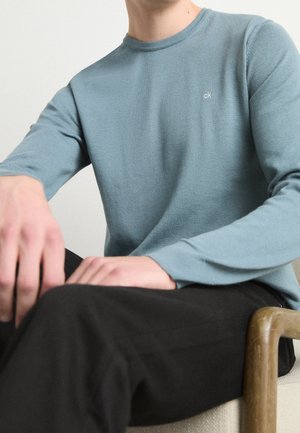 Personne portant une chemise à manches longues bleu clair avec le logo "ck" sur la poitrine, assise les mains reposant sur un pantalon foncé, un accoudoir en bois visible.