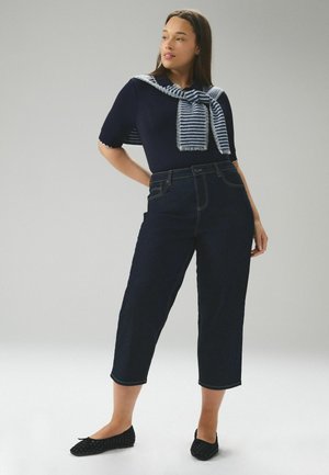 Young woman standing, wearing a navy short-sleeve top with a striped sweater draped over shoulders, dark cropped jeans, and black flats.