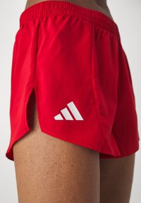 Shorts de sport rouges avec une fente latérale portés par une personne, avec un logo blanc à trois bandes près de l'ourlet.