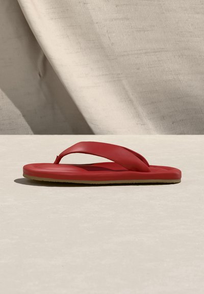 Czerwona klapka na piance z grubymi paskami, spoczywająca na jasnym kamiennym podłożu na tle beżowej tkaniny.