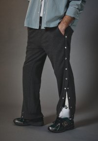 Graue Twill-Hosen mit seitlichen Druckknopfverschlüssen, versehen mit einem kontrastierenden Innenfutter. Kombiniert mit schwarzen und grünen Sneakers.