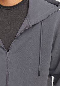Veste à zip grise avec une capuche spacieuse, fermeture éclair à l'avant et cordons de serrage. Portée par-dessus un T-shirt gris foncé, fabriqué en tissu doux.