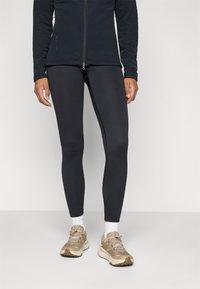 Schwarze Zip-Jacke aus strukturiertem Stoff, kombiniert mit enganliegenden schwarzen Leggings und beigen Sneakers mit weißen Socken.