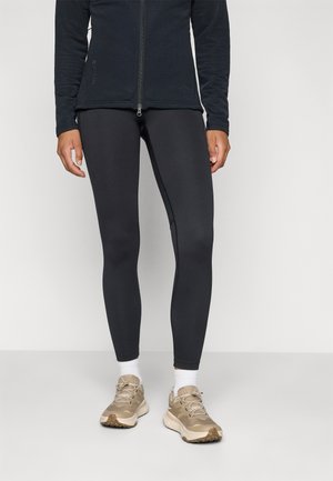 Black zip-up jacket made of textured fabric, paired with fitted black leggings and beige sneakers with white socks.
