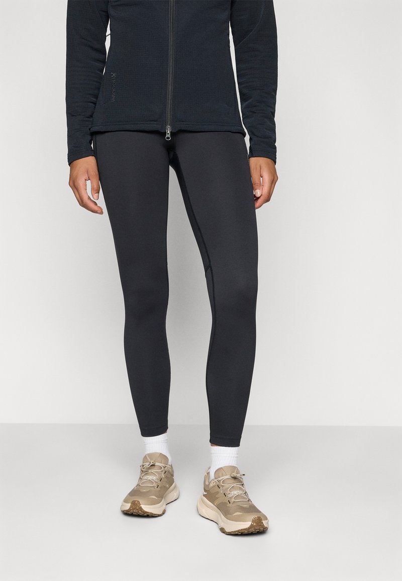 Schwarze Zip-Jacke aus strukturiertem Stoff, kombiniert mit enganliegenden schwarzen Leggings und beigen Sneakers mit weißen Socken.