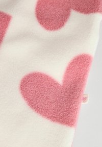 Plaid en polaire avec de grands motifs de cœurs roses sur un fond crème. Texture douce avec un léger éclat. Petite étiquette visible sur le bord.