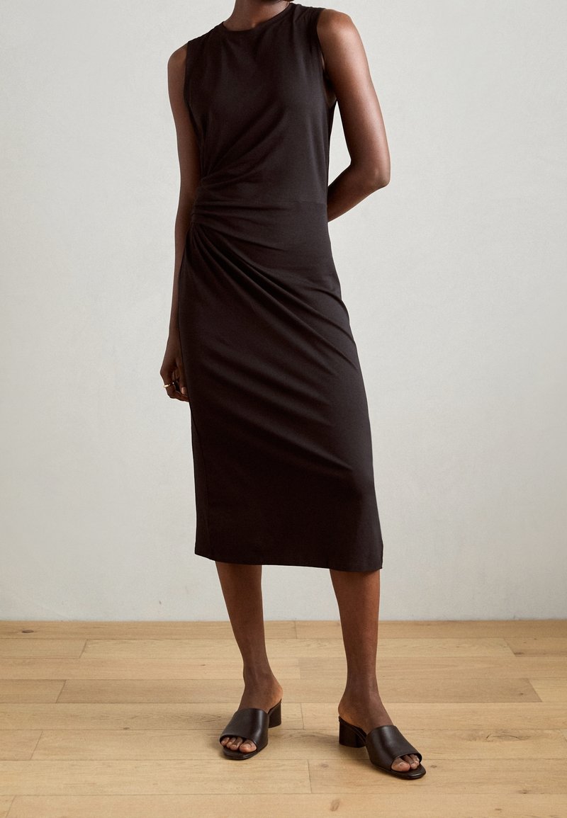 Personne portant une robe midi noire sans manches avec des fronces sur les côtés et des sandales à talons noires, debout sur un sol en bois face à un mur uni.