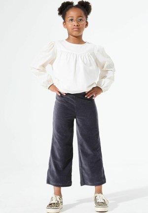 Blouse blanche à manches longues avec encolure froncée, associée à un pantalon large en velours côtelé gris foncé et des baskets à motifs noir et blanc.