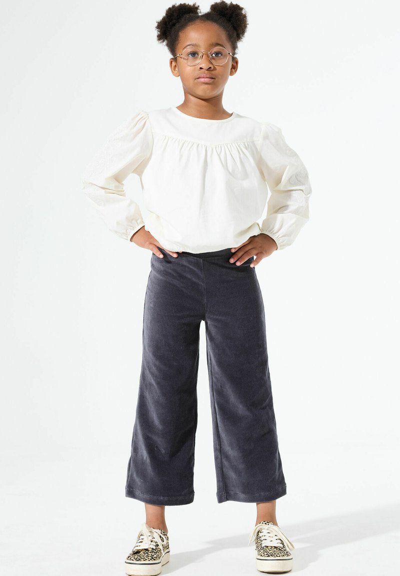 Witte blouse met lange mouwen en een geplooide halslijn, gecombineerd met donkergrijze corduroy wijde broek en zwart-wit geblokte sneakers.