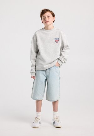 Jonge persoon staand, gekleed in een lichtgrijze sweatshirt met Los Angeles-embleem, lichtblauwe denim kniebroek, witte sokken en witte sneakers.