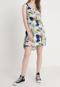 Květinové bezrukávové šaty s růžovými a modrými květy na bílém pozadí, s elastickým pasem pro uvolněný střih. V kombinaci s černými teniskami.