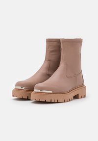 Botas de tobillo de cuero marrón con un acabado texturizado, paneles laterales elásticos y una suela de goma de gran tamaño. Presentan un acento metálico en la punta.