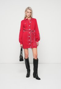 Czerwona dzianinowa sukienka z długimi puff sleeves, zapinana na guziki z przodu i związywana paskiem. Posiada wielokolorową haftę i kontrastujące białe paski. Czarne kozaki za kolano.