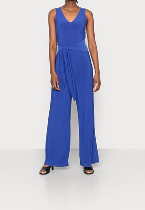 Donna che indossa una tuta royal blue senza maniche con scollatura a V profonda e una cintura annodata in vita, abbinata a tacchi neri open toe.