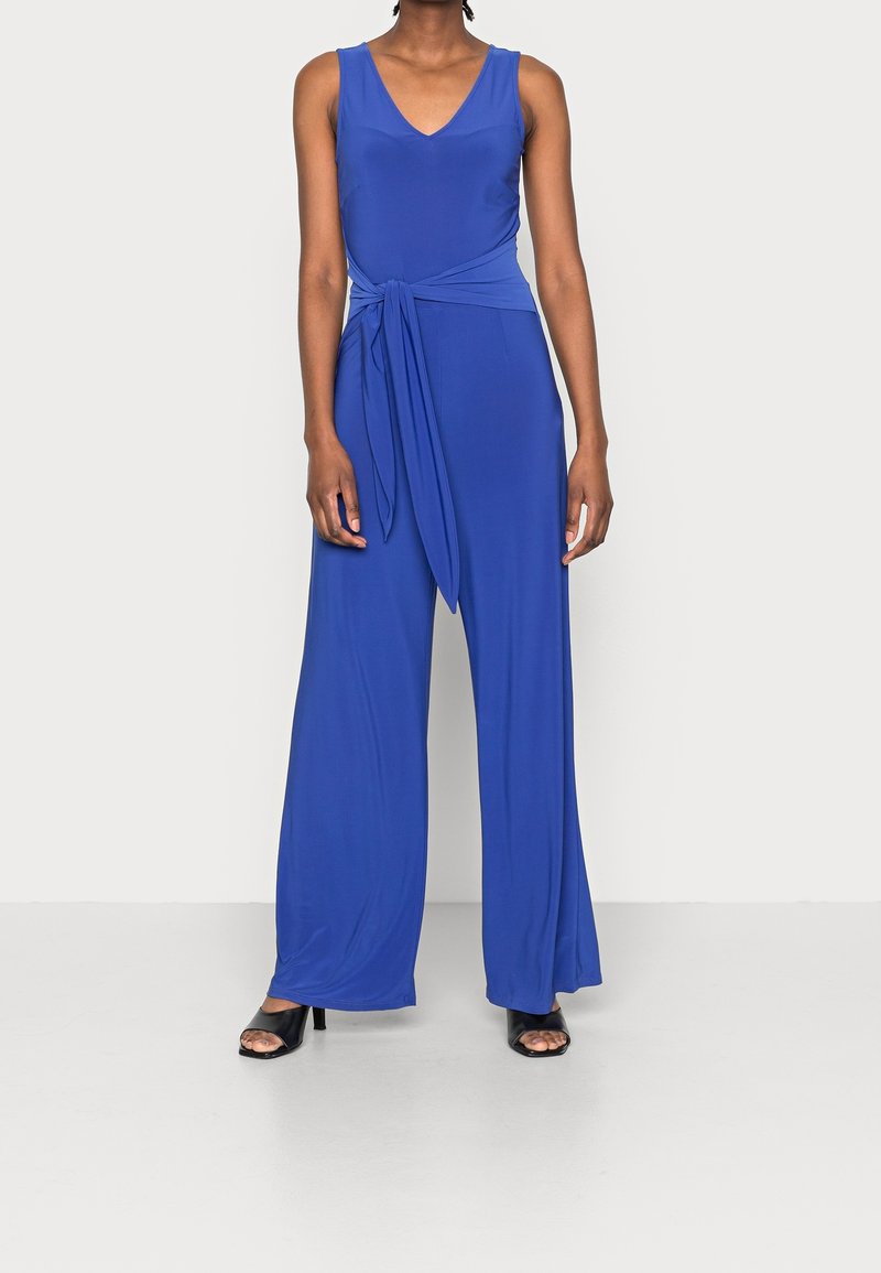 Femme portant une combinaison sans manches bleu roi avec un profond décolleté en V et une ceinture nouée à la taille, associée à des talons ouverts noirs.