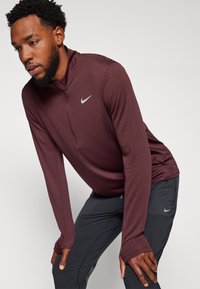 Bordo Nike majica dugih rukava s polu-zip ovratnikom, izrađena od glatkog, rastezljivog materijala. Ima otvore za palčeve i suptilni logotip na prsima.