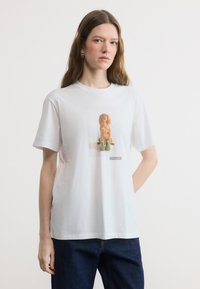 Γυναίκα που φοράει λευκό μπλουζάκι με εκτύπωση ενός καφέ σκύλου, καθισμένη πίσω από πράσινες μπότες και σκουρόχρωμο μπλε τζιν, με φόντο ένα απλό υπόβαθρο.