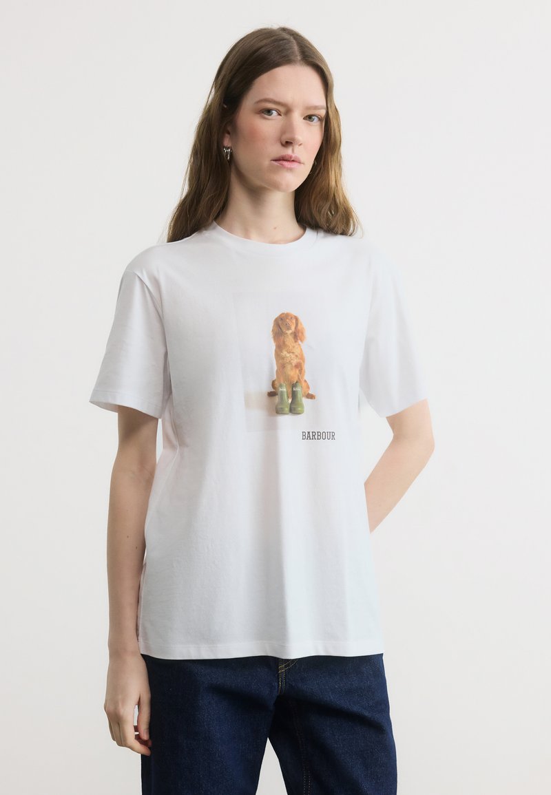 Γυναίκα που φοράει λευκό μπλουζάκι με εκτύπωση ενός καφέ σκύλου, καθισμένη πίσω από πράσινες μπότες και σκουρόχρωμο μπλε τζιν, με φόντο ένα απλό υπόβαθρο.