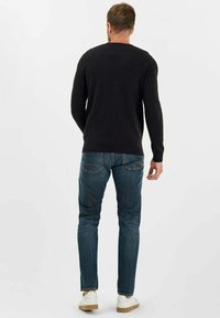 Uomo in piedi, di spalle, che indossa un maglione nero a maniche lunghe, jeans blu e sneakers bianche su uno sfondo bianco.