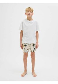 T-shirt blanche à manches courtes associée à un short beige à motifs avec des feuilles vertes et des fleurs oranges. Debout pieds nus, fond neutre.