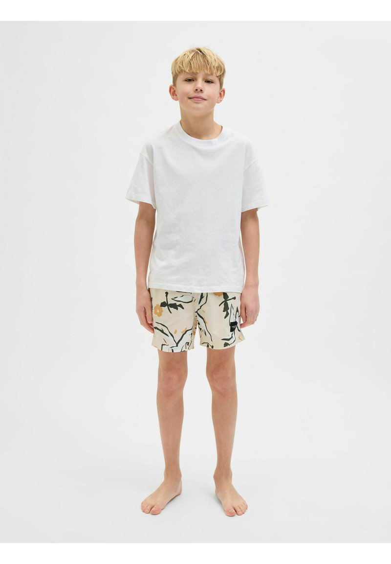 T-shirt blanche à manches courtes associée à un short beige à motifs avec des feuilles vertes et des fleurs oranges. Debout pieds nus, fond neutre.