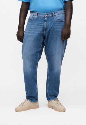 Mand iført løse blå jeans, beige sneakers og en blå skjorte, der står mod en ensfarvet hvid baggrund.