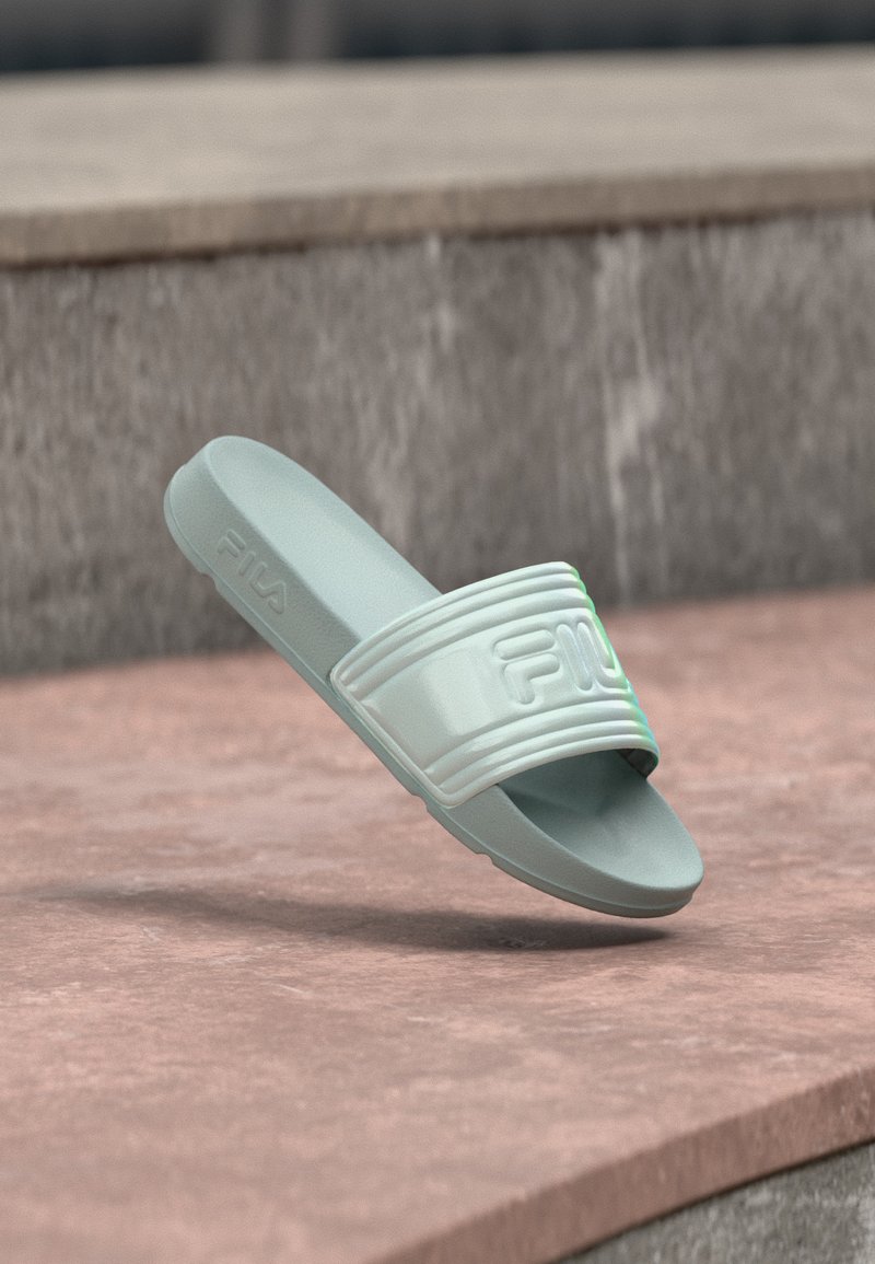 Ανοιχτό γκρι σαγιονάρα Fila slide με ανάγλυφο λογότυπο στο λουράκι, ανυψωμένη πάνω από μια πέτρινη επιφάνεια και φόντο από σκυρόδεμα.