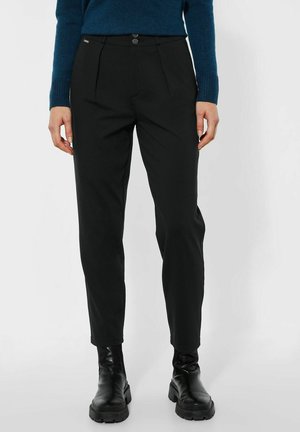 Personne portant un pantalon noir fuselé, des bottines noires épaisses et un pull bleu à manches longues, debout contre un fond uni.